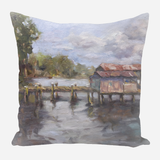 Painterly landscape on pillow of a weathered dock with a rustic building over calm water under a cloudy sky and surrounding green trees.
