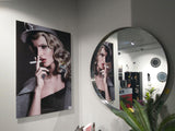 Portrait of a woman with curled blonde hair and red lipstick smoking a cigarette, wearing a gray coat and hat, reflected in a large round wall mirror.
