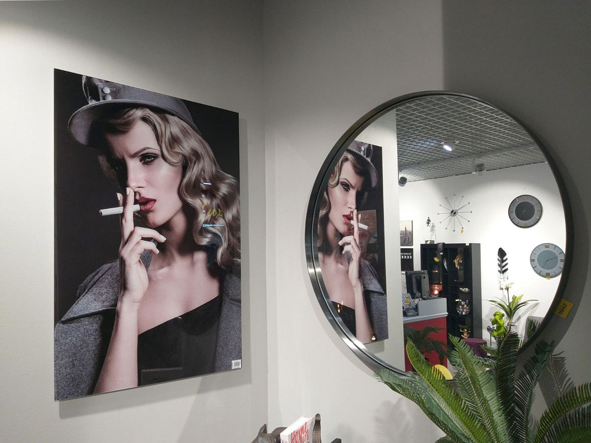 Portrait of a woman with curled blonde hair and red lipstick smoking a cigarette, wearing a gray coat and hat, reflected in a large round wall mirror.
