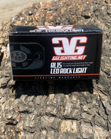 Black rectangular box labeled GGLighting.net RL15 LED Rock Light with red and white text, placed on textured rough brown rock surface outdoors.