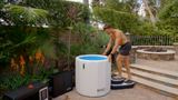 Man stepping onto black inflatable steps beside a white inflatable barrel with blue interior, set outdoors near stone steps and lush greenery.