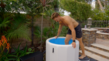 Shirtless man in dark swim trunks stepping into a white and blue inflatable barrel pool in a backyard with tropical plants and stone steps.