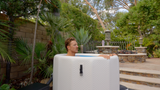 Person relaxing in a white, square inflatable barrel with textured sides and black handles, set outdoors near palm trees and stone steps.