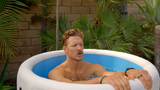 Man relaxing with eyes closed in a white and blue inflatable hot tub surrounded by tropical plants and a stone wall.
