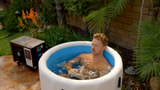 Man relaxing in a round blue inflatable barrel with white rim, surrounded by green plants and a stone floor in a backyard setting.