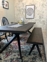 Rectangular dining table with dark wood slab top and black angled metal legs, paired with a matching wooden bench and black chairs on a multicolored patterned rug.