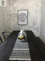 Rectangular dark wood dining table with black and white geometric patterned runner and a fruit bowl holding bananas, oranges, and a lime under a framed tree ring print on textured white wall.
