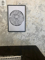 Black and white framed print of a detailed Douglas Fir tree cross-section hung on a textured light gray and beige wall above a dark wood table with a fringed black and white textile edge.