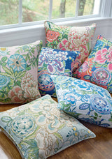 Six decorative pillows featuring detailed floral patterns in pastel and vibrant shades, arranged on a wooden surface near a window with a natural outdoor backdrop.