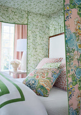 Bedroom corner with a white bed featuring a green-striped duvet, floral Peony Garden pillow with blush background, and a pink pillow with geometric pattern, light green leafy canopy overhead.