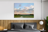 Canvas painting above bed showing expansive green field under a vibrant sky with clouds in shades of blue, peach, and white, framed by dark tree silhouettes on horizon.
