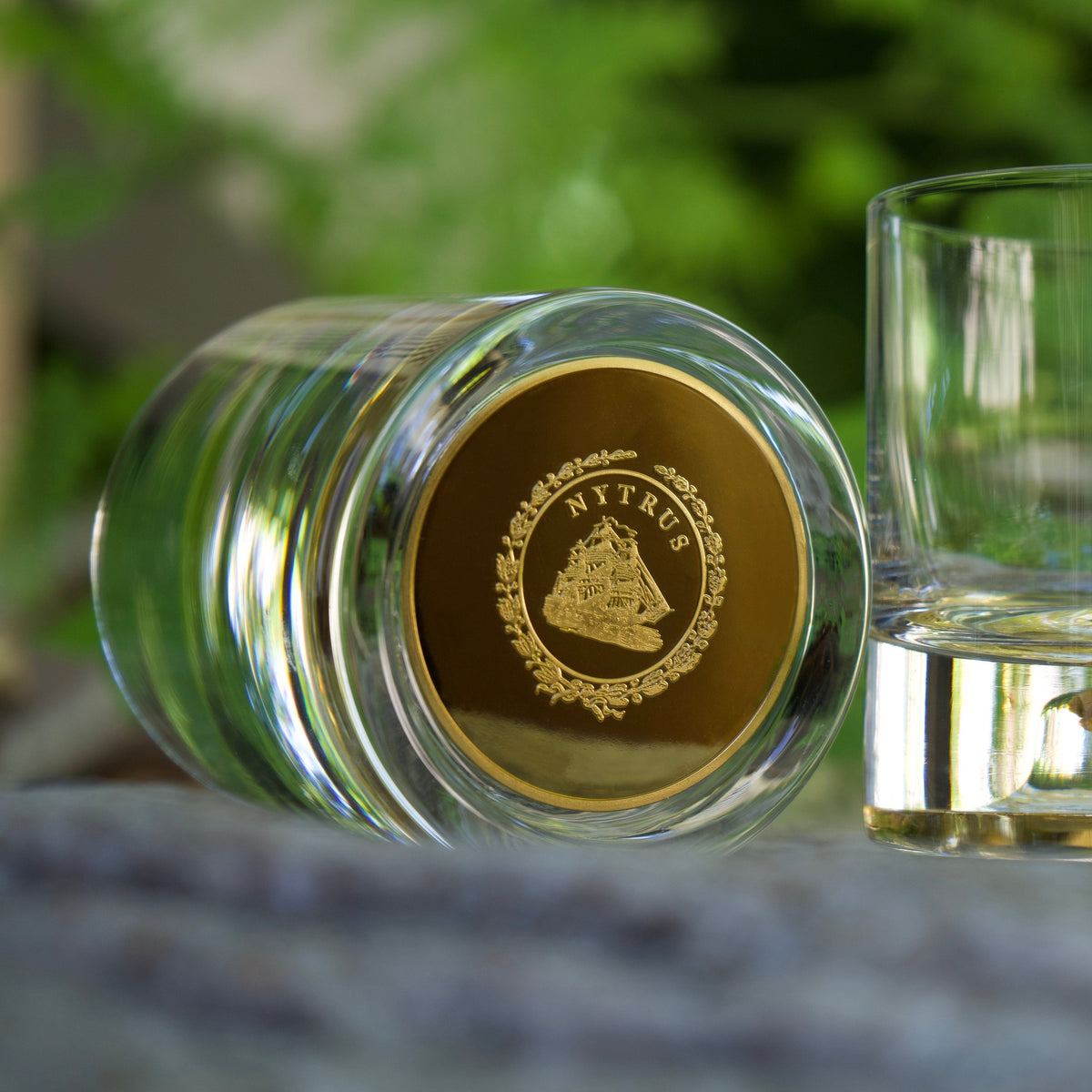 Close-up of a clear glass tumbler lying on its side, featuring a gold medallion inset at the bottom with an intricate emblem and lettering.
