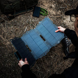 Person holding an unfolded blue foldable solar panel outdoors on dry ground next to a green paracord coil and a backpack, viewed from above and slightly behind.