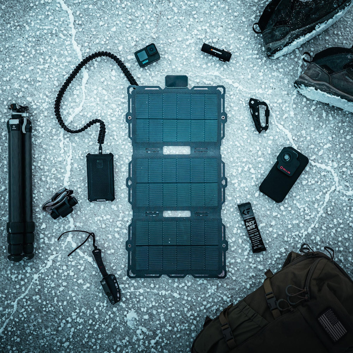 Folded black solar panel on speckled gray surface surrounded by camera gear, hiking boots, and brown backpack, arranged in a flat lay composition.