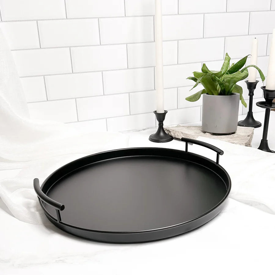 Black round tray with handles on a white surface with a plant and candles in the background.