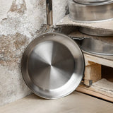 Round stainless steel frying pan with a long handle resting against a textured stone wall and wooden shelf with stacked metal cookware.