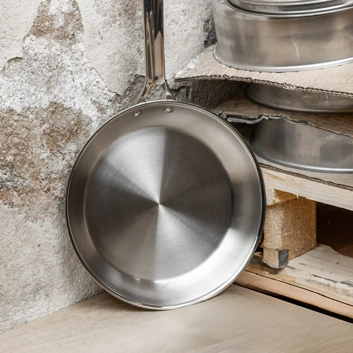 Round stainless steel frying pan with a long handle resting against a textured stone wall and wooden shelf with stacked metal cookware.