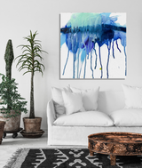 Abstract canvas print featuring blue and teal paint dripping vertically on a white background, displayed above a white sofa in a minimalist room with potted plants and a round wooden coffee table.