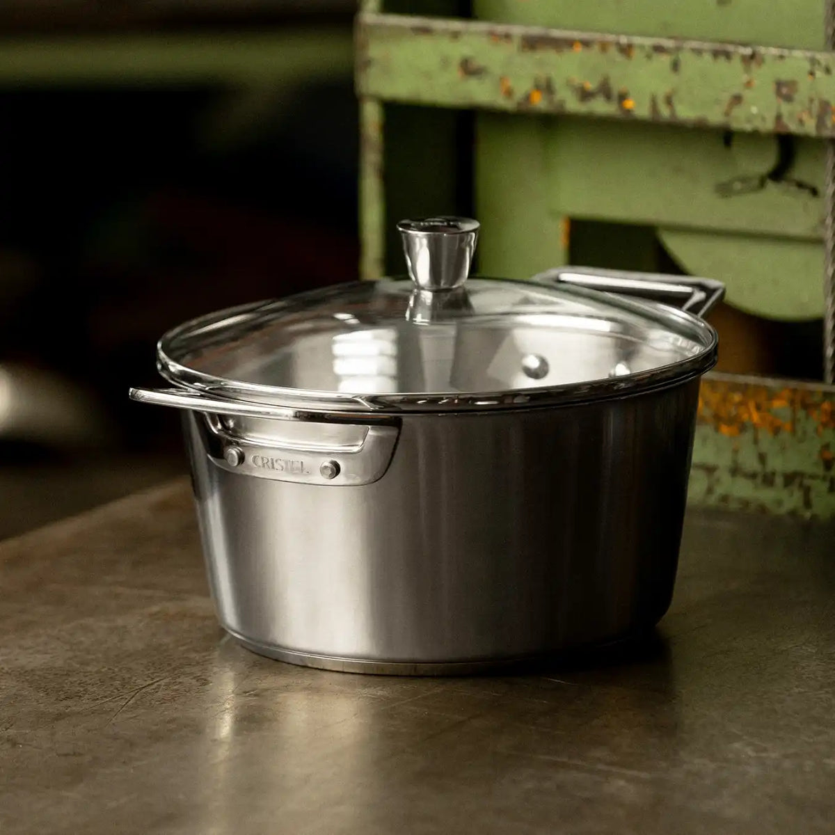 Stainless steel stewpot with two short handles and a transparent glass lid with a metal knob, placed on a worn metal surface near a green rustic structure.