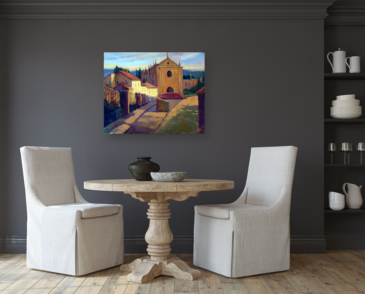Painting of a sunlit Portuguese street featuring a stone church and houses with warm yellow and brown tones, displayed above a round wooden table and two beige upholstered chairs.