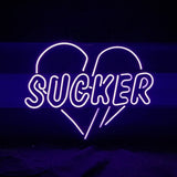 Purple neon sign displaying the word 'SUCKER' with the letters enclosed inside a stylized heart outline on a black background.