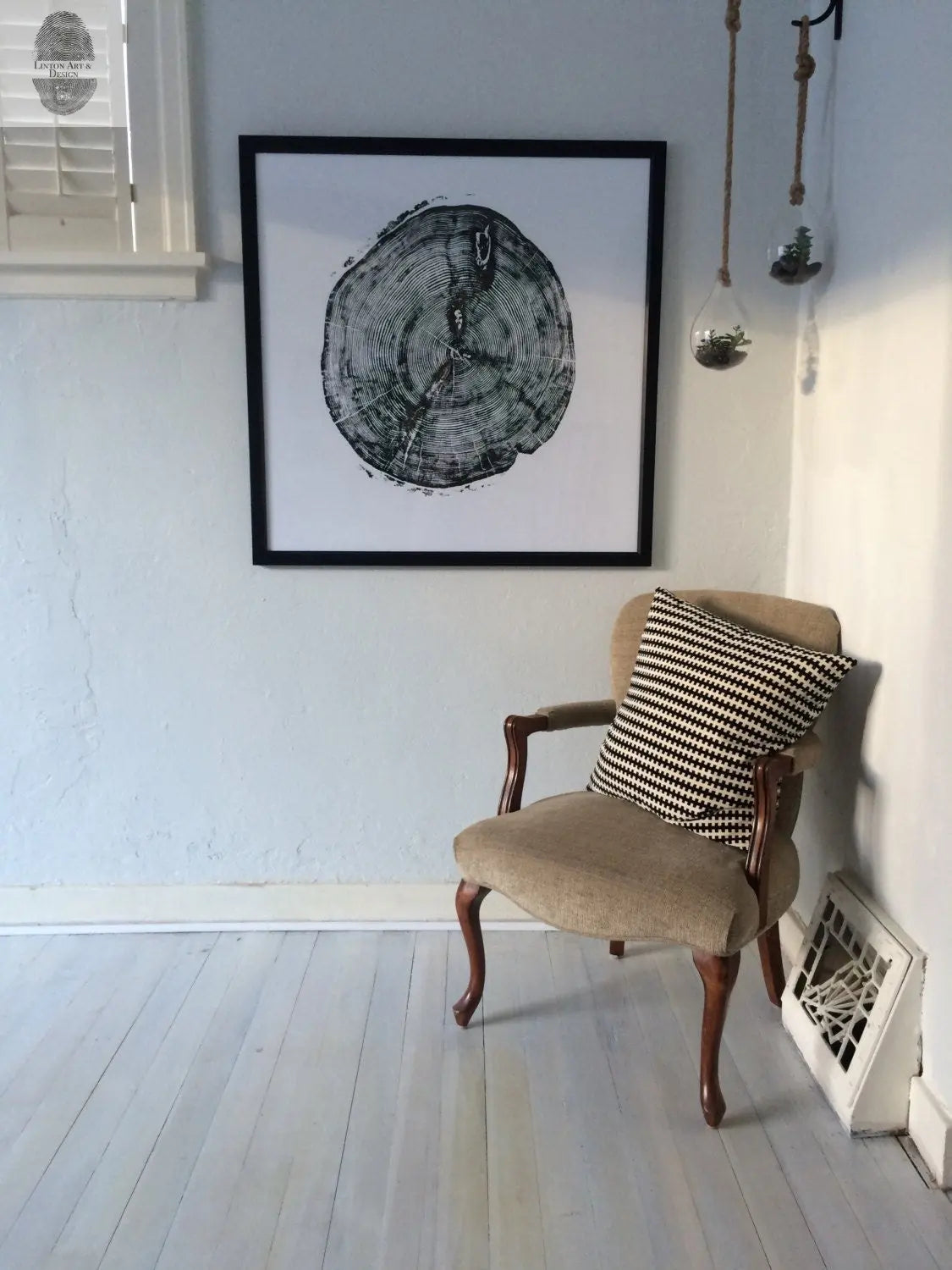 Black and white framed print of a white pine tree cross-section hanging on a pale wall above a beige upholstered chair with wooden legs and a black-and-white houndstooth pillow.