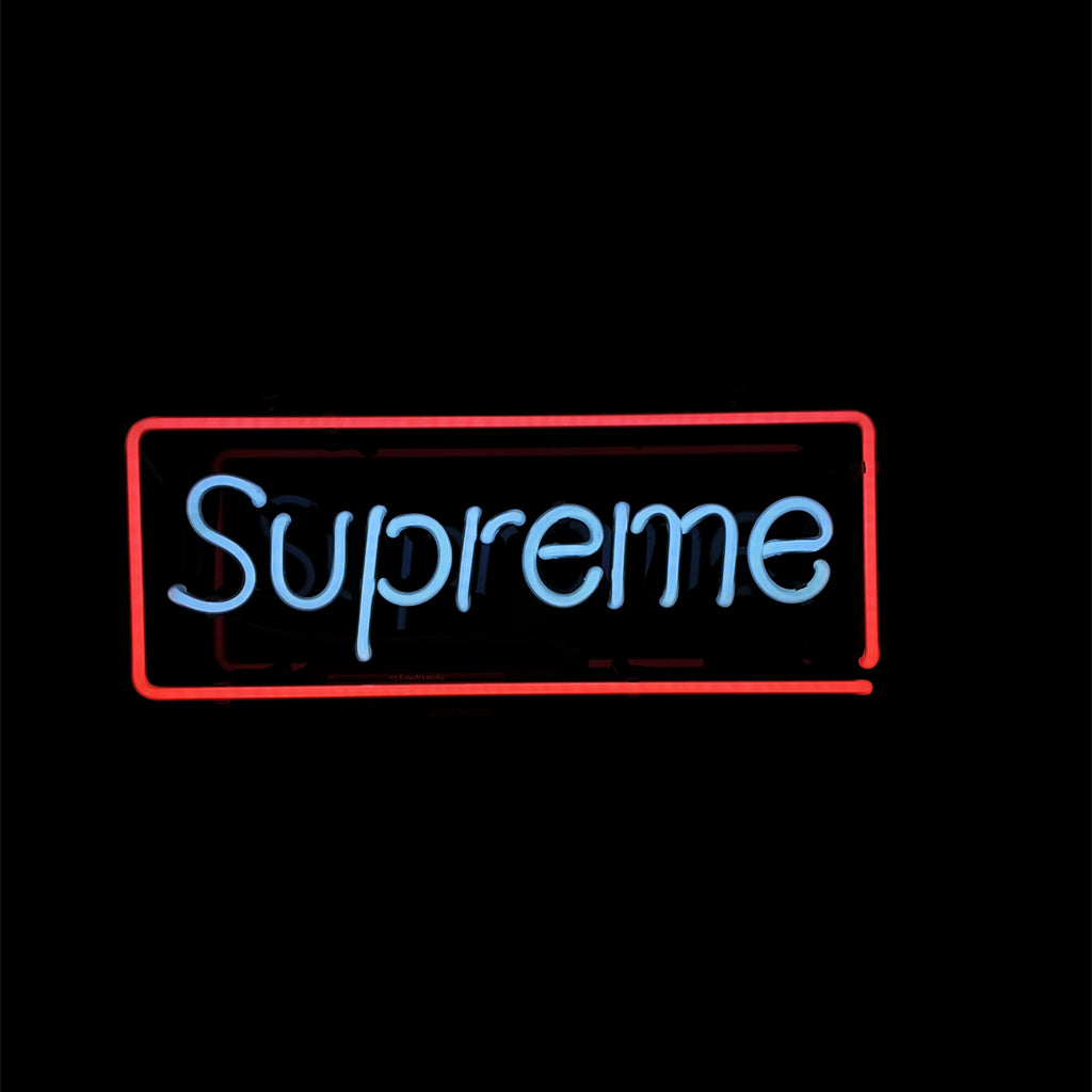 Neon sign with the word 'Supreme' in light blue script, surrounded by a rectangular red neon border on a black background.