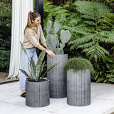 Woman adjusting tall ribbed grey planter holding a large cactus, flanked by two shorter ribbed grey planters with snake plant and bushy foliage, set on white tile patio.