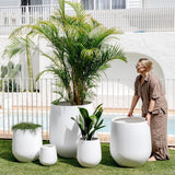 Five smooth, white ceramic planters of varying sizes arranged on green grass by a pool, with a person adjusting the largest planter on the right.