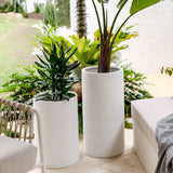 Two tall white cylindrical ceramic planters with green leafy plants, placed on a light outdoor patio near wicker and cushioned seating with a striped pillow.