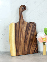 Large rectangular wooden cutting board with a curved handle featuring a hole, showing dark and light natural wood grain patterns, standing upright on a marble surface.