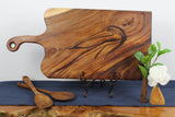 Rectangular wood board with a handle and etched pelican design, displayed on a decorative stand alongside wooden spoons and a small vase with green leaves and a white flower.