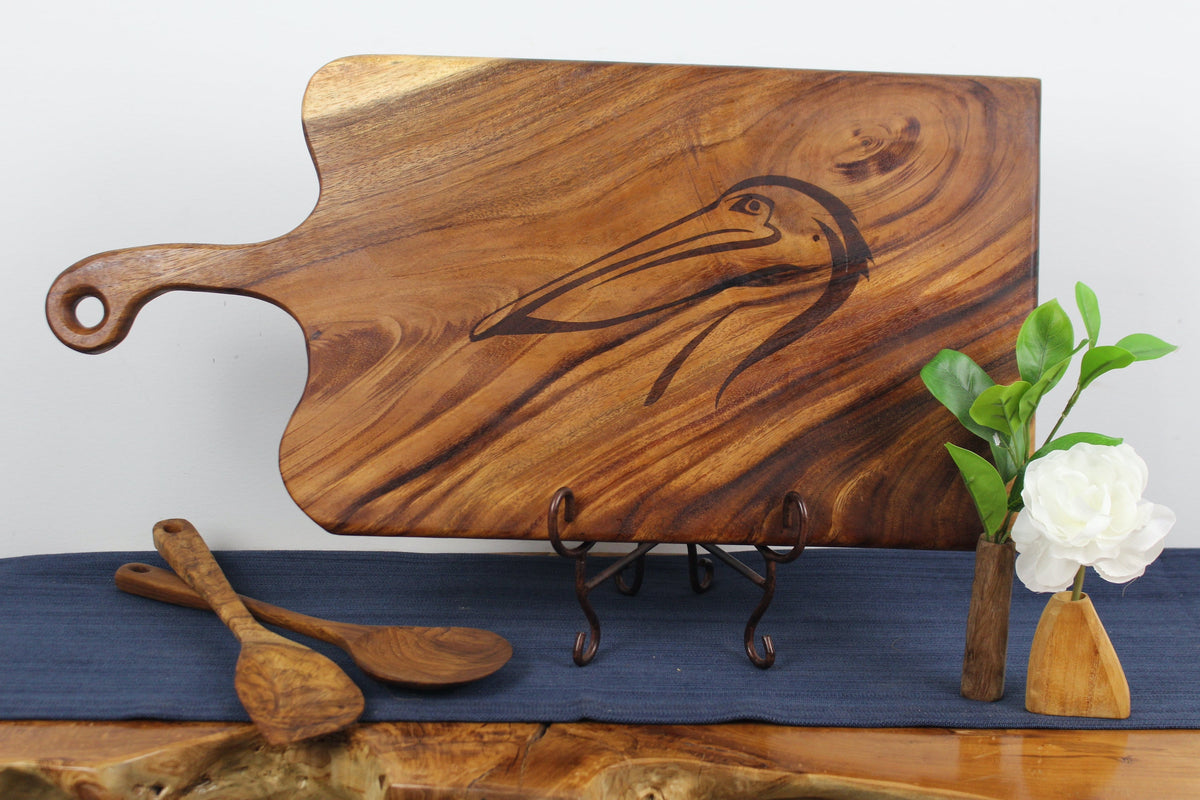 Rectangular wood board with a handle and etched pelican design, displayed on a decorative stand alongside wooden spoons and a small vase with green leaves and a white flower.