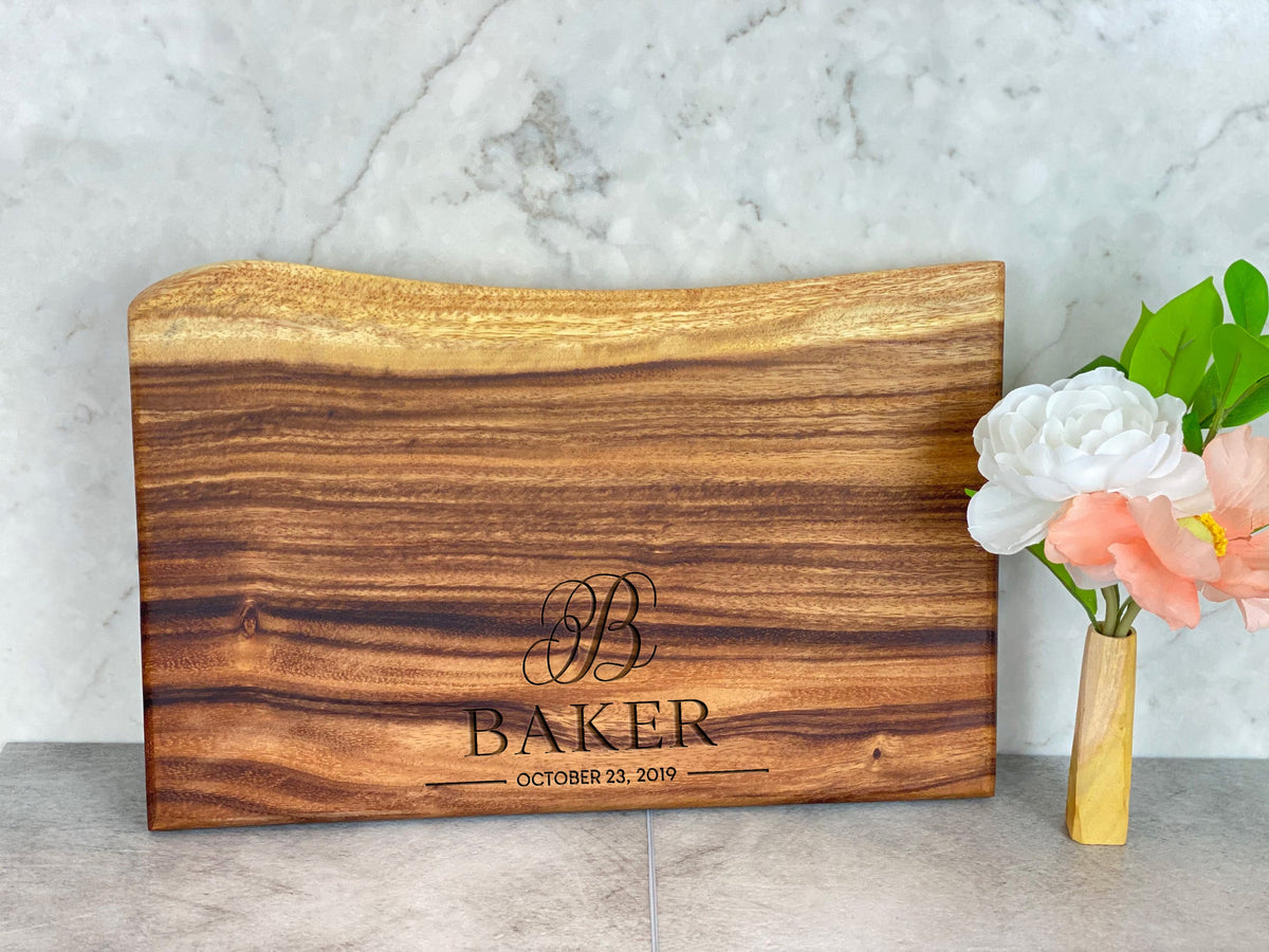 Medium live edge cutting board with a smooth square end, dark and light brown wood grain, and engraved text reading BAKER October 23, 2019 on a marble surface.