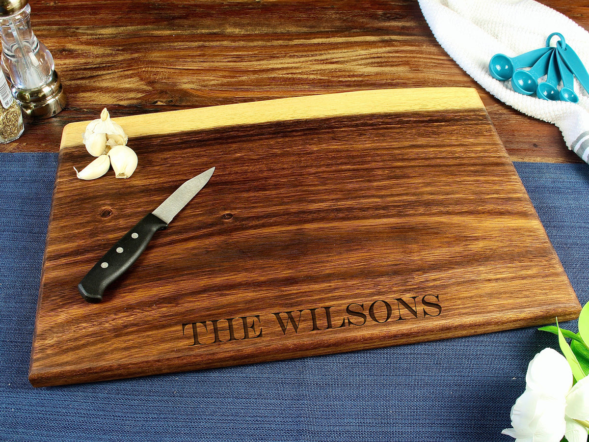 Rectangular dark wood live edge cutting board with engraved text 'THE WILSONS' near edge, shown with a small black-handled knife and garlic cloves on top.