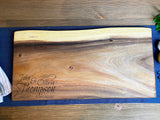 Large rectangular live edge wood board with natural grain, dark brown upper band, and engraved script names and date on bottom left corner, displayed on a blue cloth.