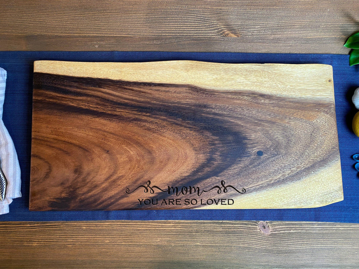 Large rectangular wooden board with natural live edges, featuring dark and light grain patterns and the phrase 'mom YOU ARE SO LOVED' engraved in black script.