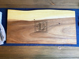 Rectangular live edge wood board with a lighter top edge and engraved monogram 'L' in the center, placed on a dark blue cloth over a wooden table.