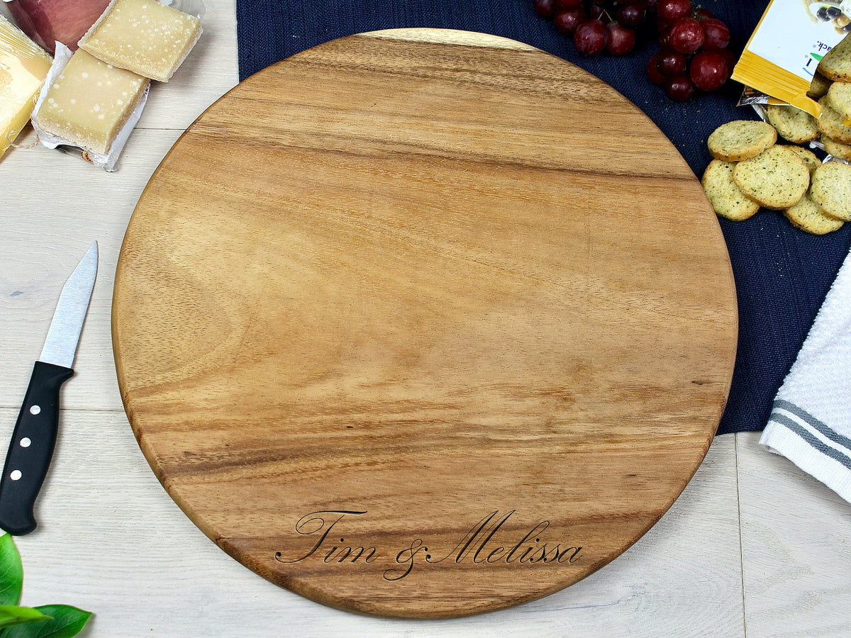 Medium round wooden charcuterie board with smooth grain and engraved "Tim & Melissa" lettering, shown surrounded by cheese, grapes, crackers, and a knife on a light wood surface.