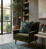Dark olive green velvet armchair with a deep blue floral patterned pillow, placed next to a modern side table and a large potted plant near floor-to-ceiling windows.