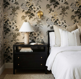 Black bedside table with two drawers, a beige lamp, clear glass bottle, and open book beside a bed with white and ivory embroidered pillows against floral wallpaper.