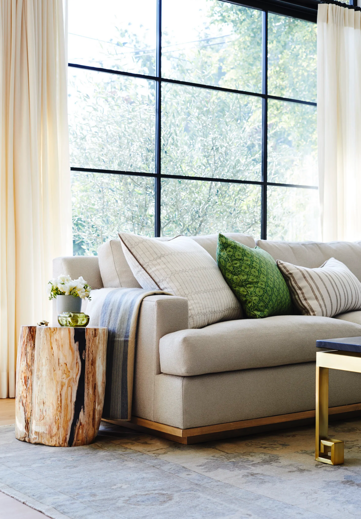 Neutral-toned sofa with textured sand-colored and green twine-patterned pillows near a large window, next to a natural wood stump side table holding a small plant.