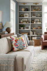 Beige sofa with a coral and yellow floral patterned pillow, positioned in front of gray built-in shelves filled with pottery and framed photos in a softly lit living room.