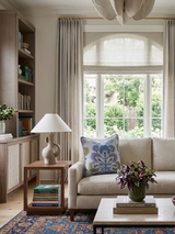 Living room corner with beige sofa featuring a blue and lavender floral pillow, a wooden side table with a sculptural lamp, and a coffee table holding a leafy plant.