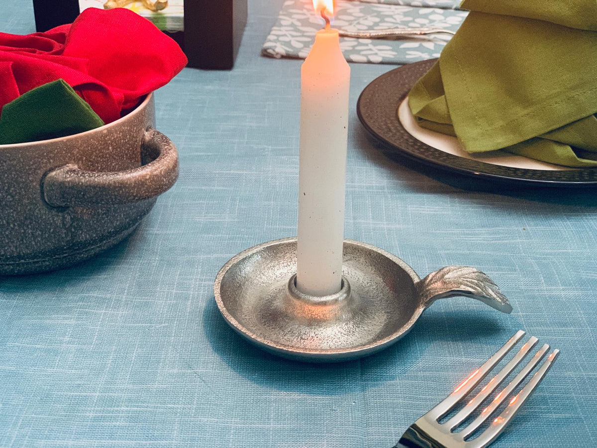 Silver-toned metal taper candle holder dish with a curved handle, holding a lit white candle on a blue textured tablecloth beside a fork and bowls with folded cloth napkins.