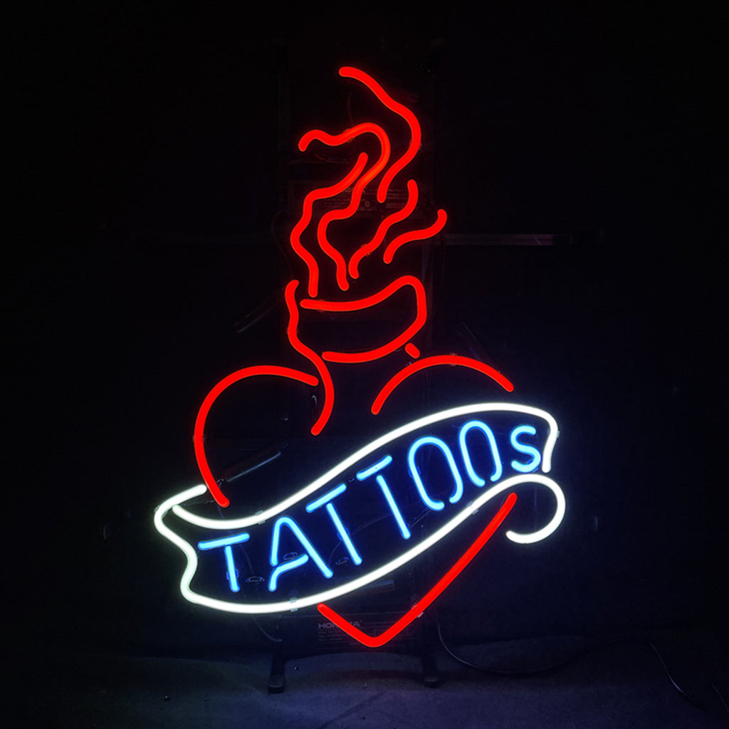 Neon sign of a red heart with flames above it and a white banner across the heart reading 'TATTOOS' in blue letters against a black background