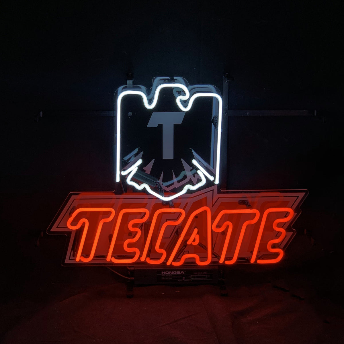 Red neon letters spelling 'TECATE' beneath a white neon outline of an eagle emblem on a black background