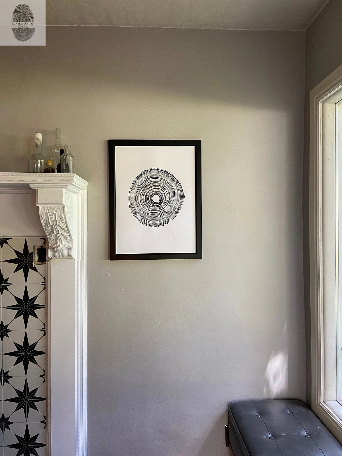Black and white circular tree ring print in a black frame hanging on a light gray wall, near a white fireplace with black star-patterned tile and a tufted gray bench by a window.