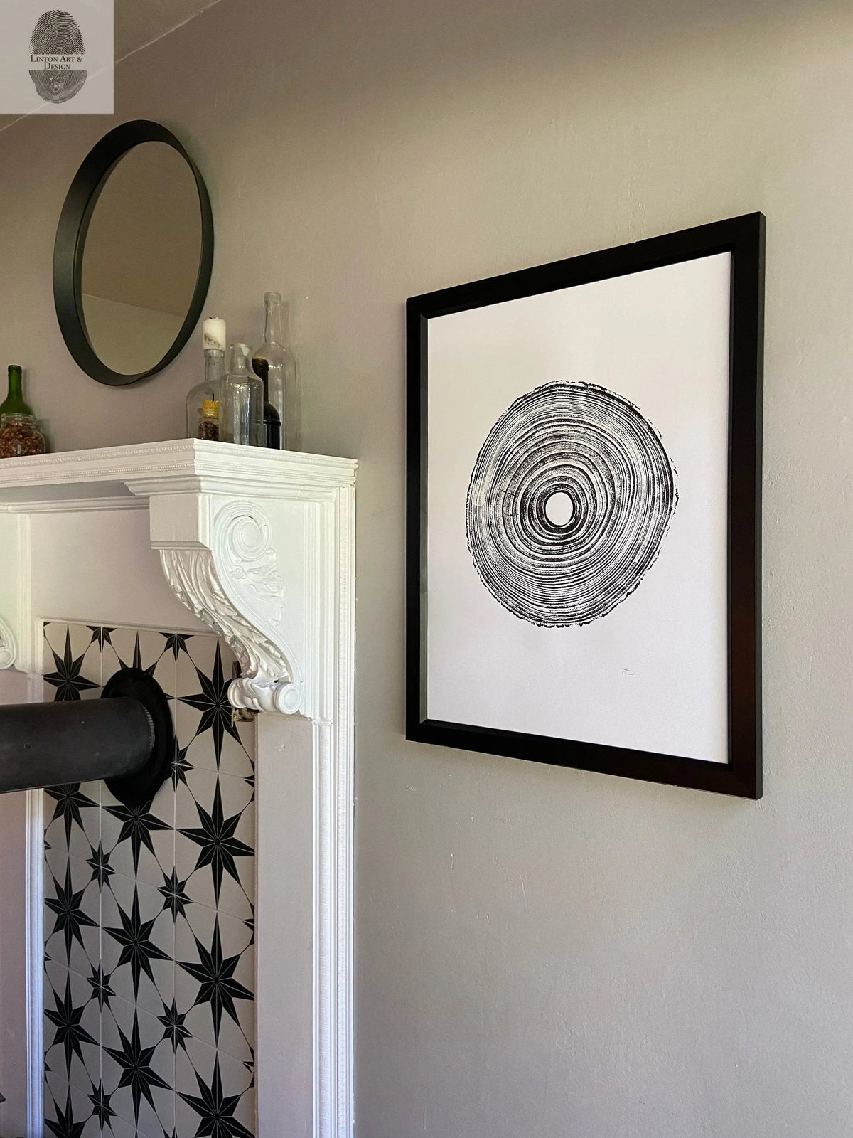 Black and white concentric tree rings print in a black frame hung on a gray wall next to white fireplace mantel with black and white star-patterned tiles.
