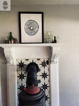 White mantel with ornate corbels above a black wood stove pipe set against black and white star-patterned tiles, topped with a black-framed tree ring print and glass bottles.